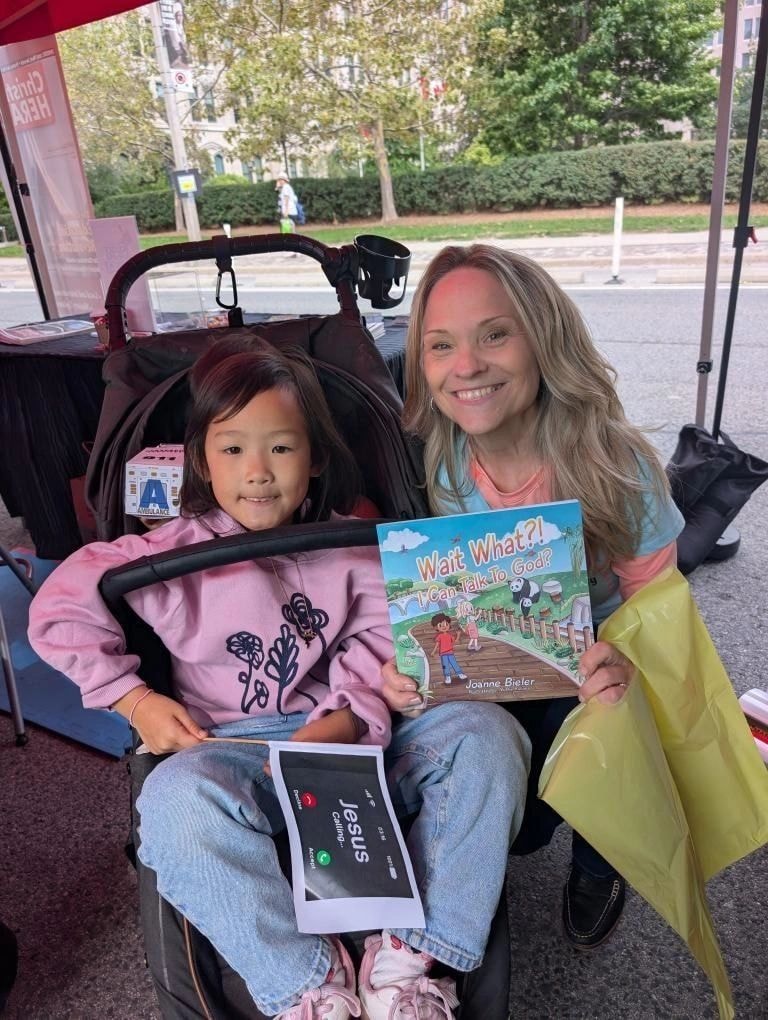 Joanne with a sweet little girl in a stroller holding Wait What?! I Can Talk To God? at JITC Parade 2025