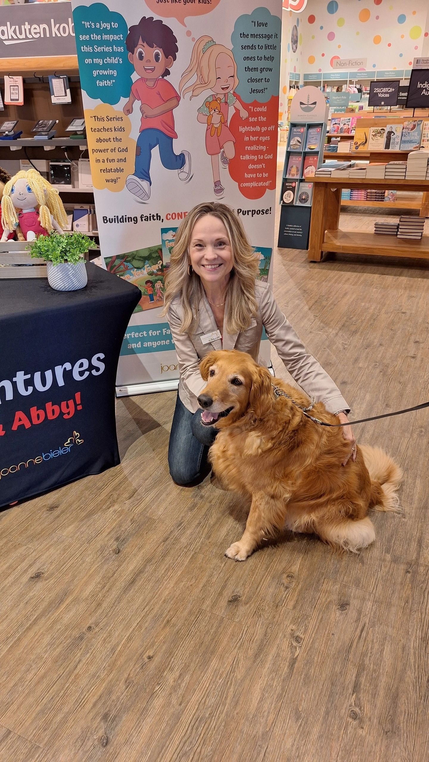Joanne kneeling with a golden retriever dog at her book signing booth at Indigo Erin Mills