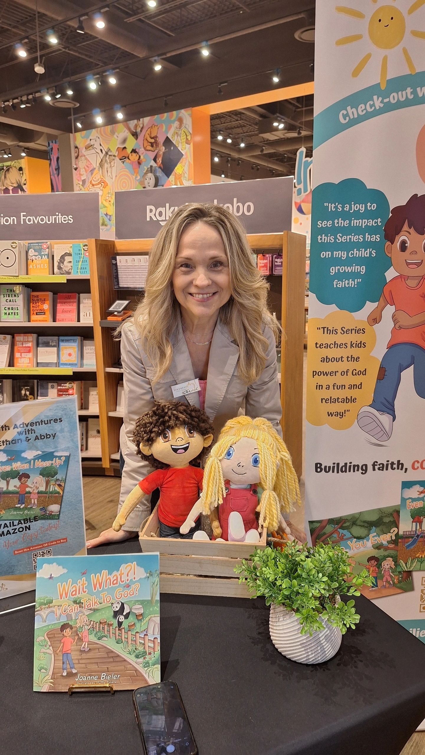 Joanne posing with Ethan and Abby plushies at her author signing table at Indigo Erin Mills