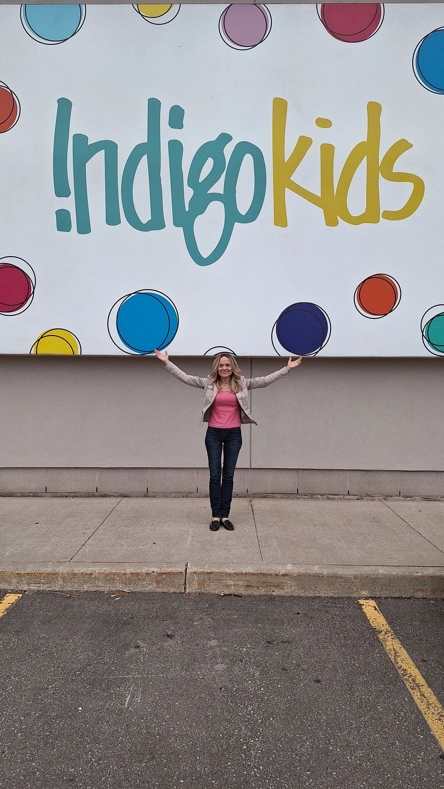 Joanne standing arms wide open in front of the Indigo Kids mural outside the store