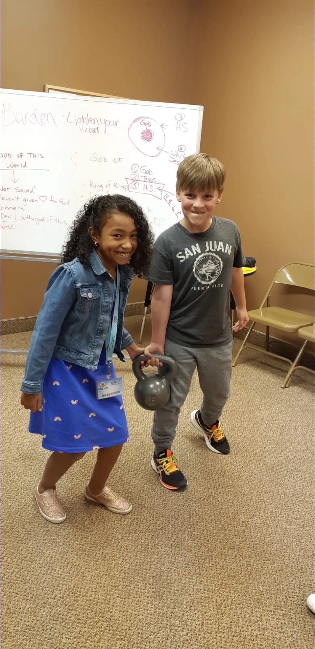 Two children holding a kettlebell together in front of a Holy Spirit lesson whiteboard