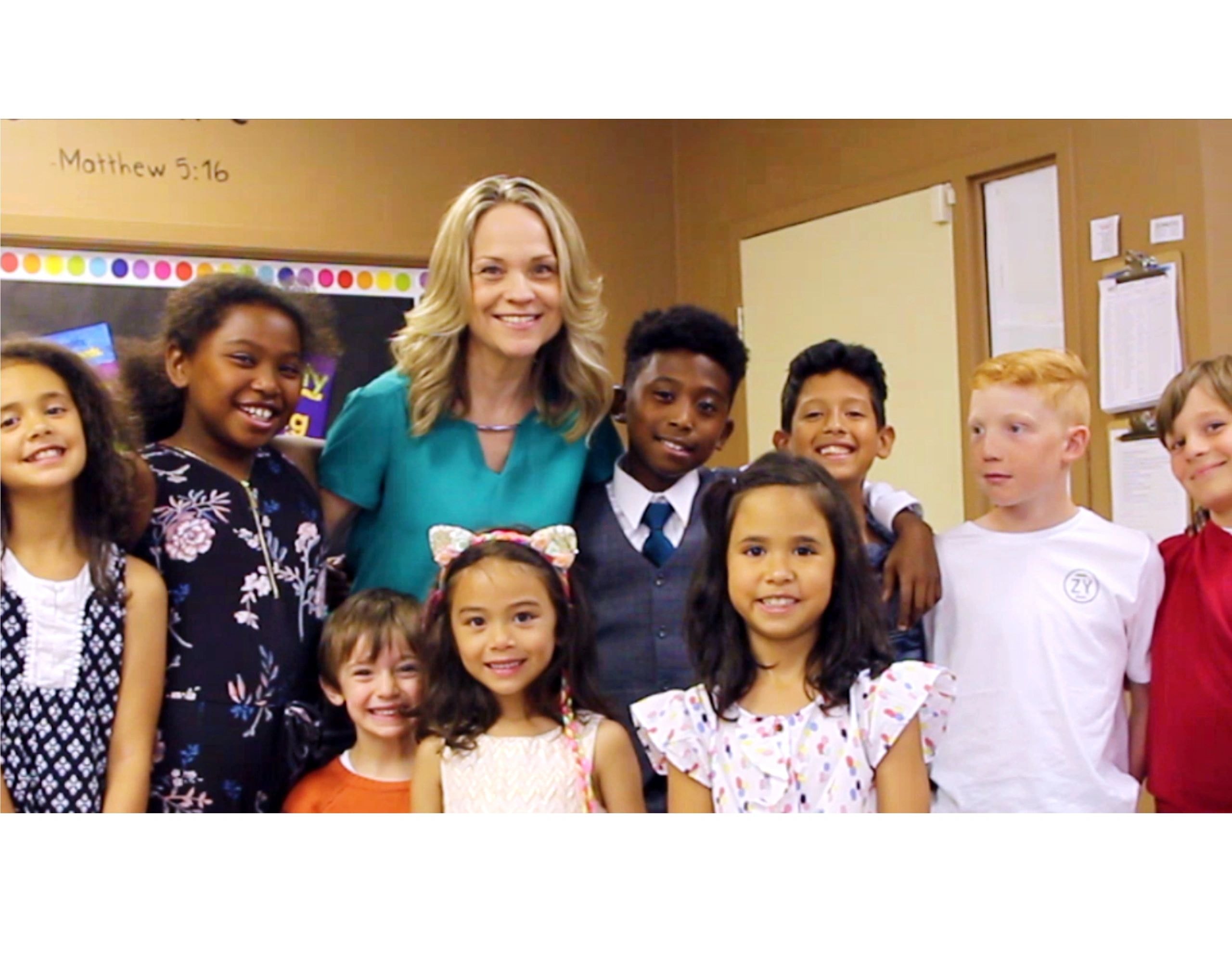 Joanne surrounded by a group of children in a classroom with Matthew 5:16 on the board