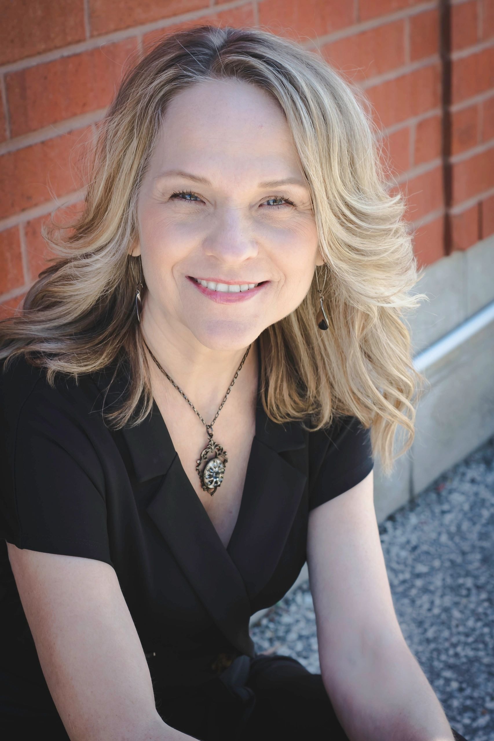 Joanne Bieler author headshot smiling against a brick wall in a black top with pendant necklace