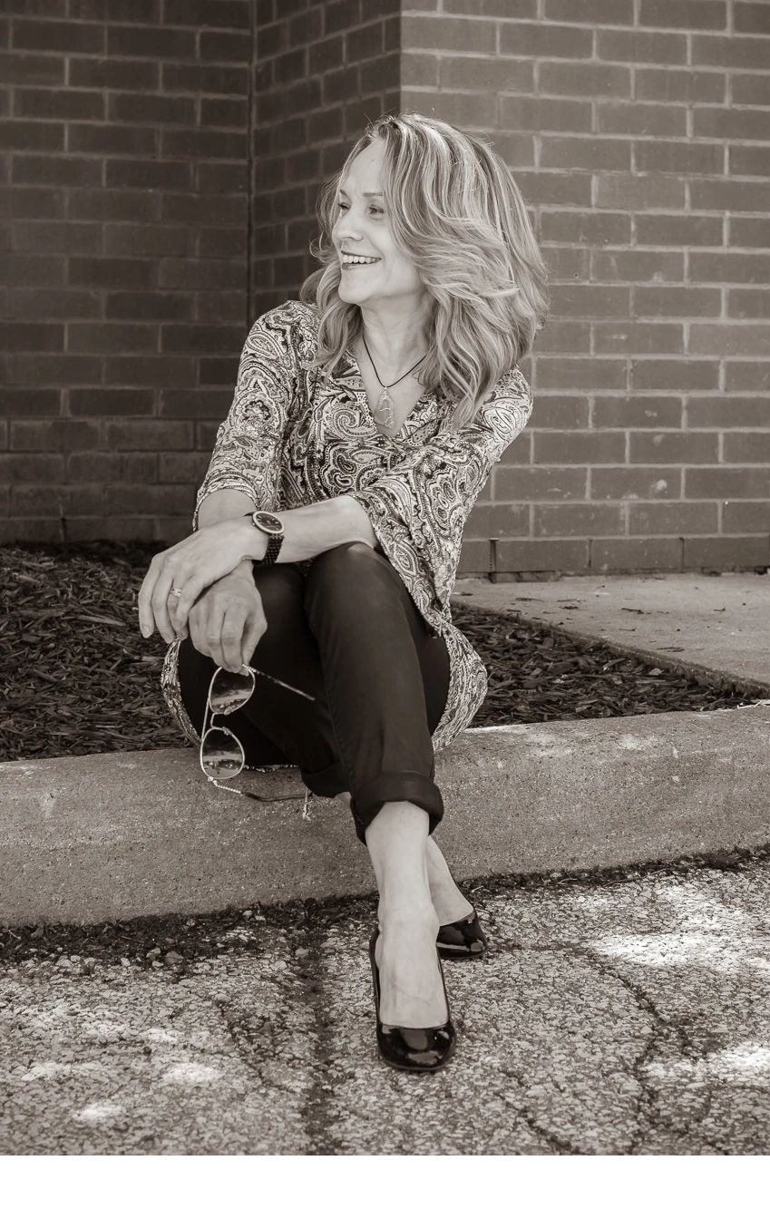 Black and white photo of Joanne sitting on a curb laughing and holding sunglasses outdoors