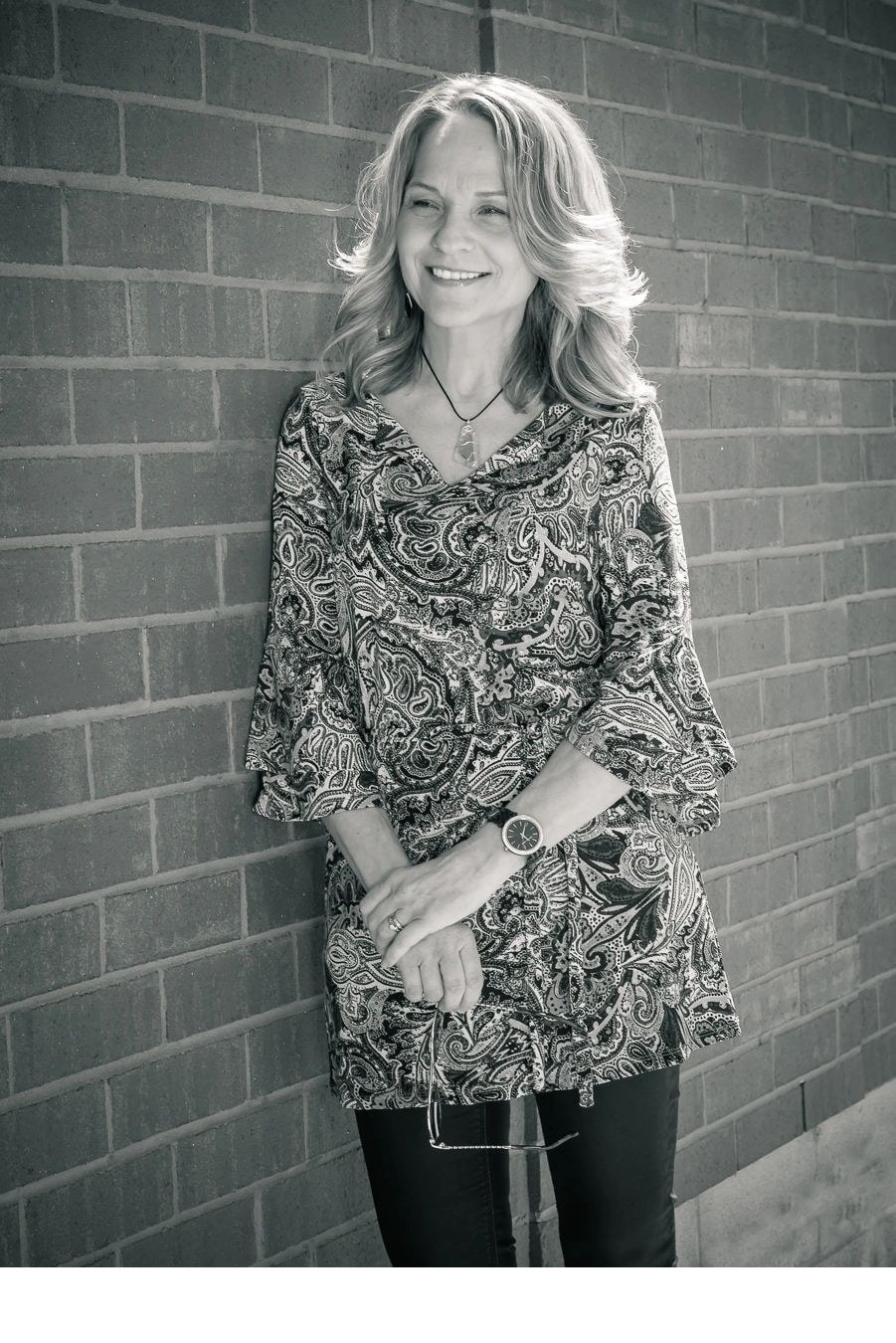 Black and white photo of Joanne leaning against a brick wall in a paisley blouse smiling
