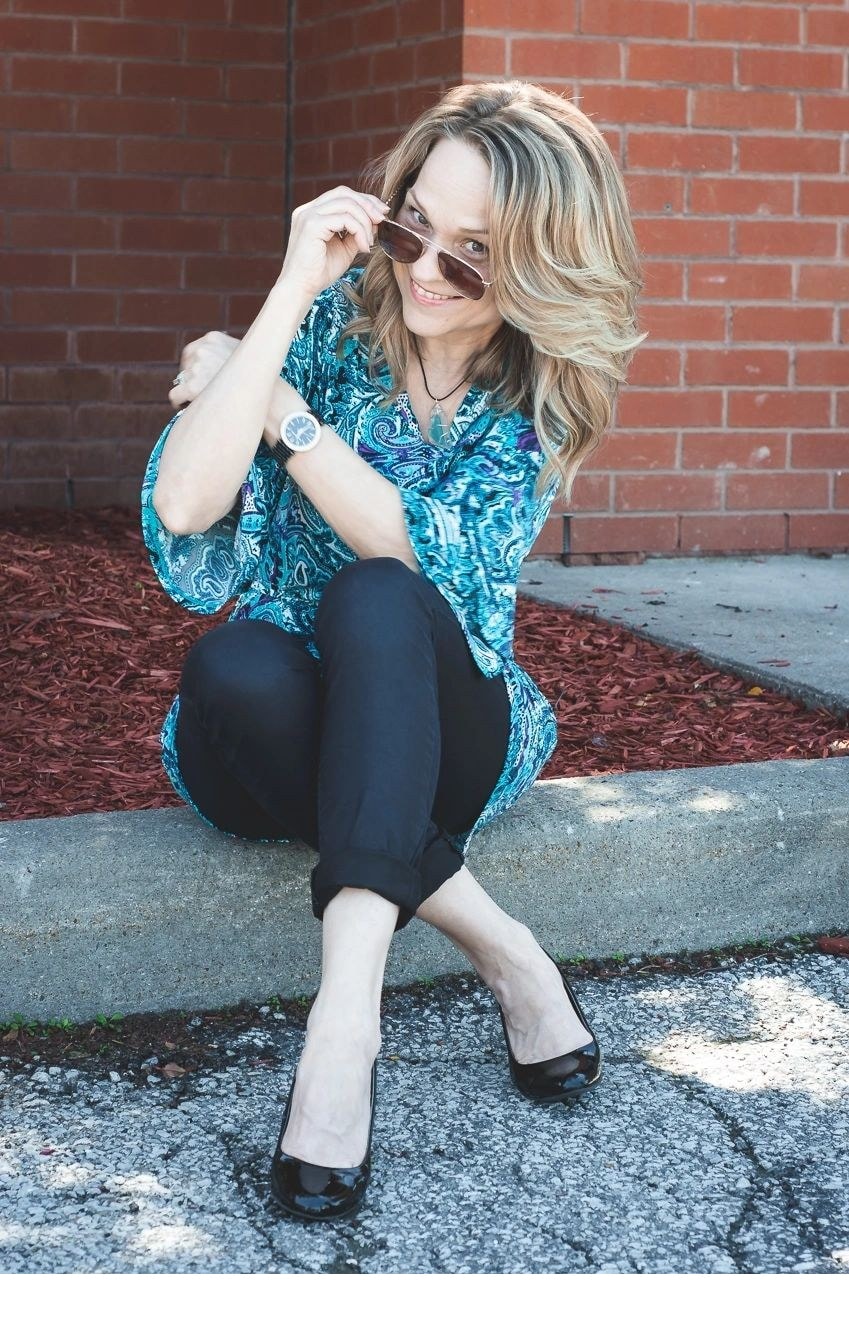 Joanne sitting on a curb in a teal paisley top holding sunglasses and smiling playfully