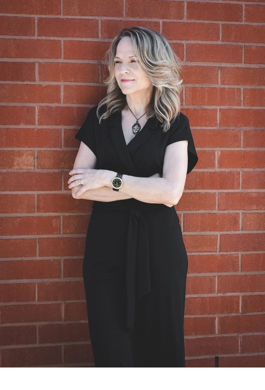Joanne standing against a brick wall in a black wrap dress with arms crossed looking confident