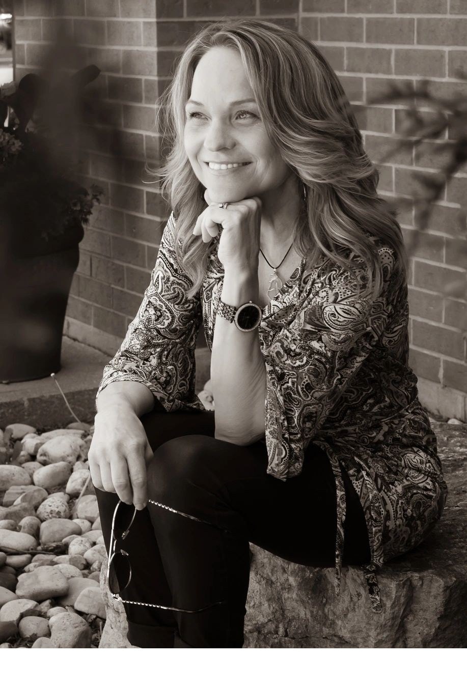 Black and white photo of Joanne sitting on a stone with chin resting on hand smiling warmly